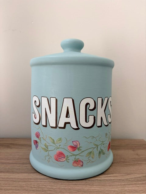 A light blue ceramic canister with a lid, featuring the word 'SNACKS' in bold white letters with dark outlines and a floral pattern near the bottom, sitting on a wooden surface against a plain beige wall.