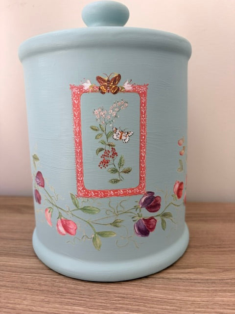 A light blue ceramic canister with a matching lid, decorated with a central floral frame design and pink berries, sitting on a wooden surface against a neutral background.