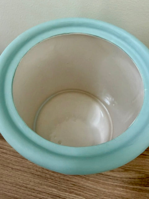 A close-up of a round ceramic container with a light blue exterior and a cream-colored interior, shown on a wooden surface against a plain wall.