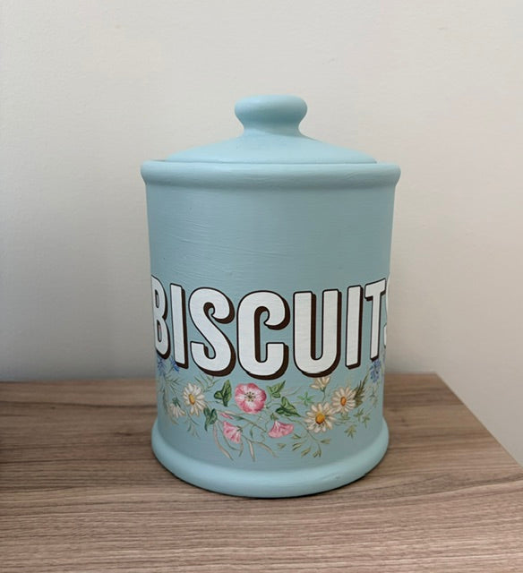 A light blue ceramic biscuit canister with a matching lid, featuring the word 'BISCUIT' in bold white letters with black outlines and a delicate floral pattern at the base, sitting on a wooden surface against a plain wall.