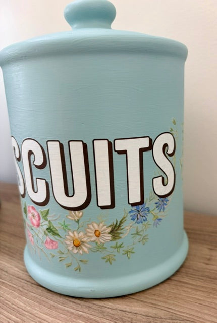 A light blue ceramic canister with a lid, decorated with the word 'BISCUITS' in large white letters and a floral design below, sitting on a wooden surface.