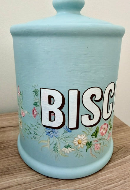 A light blue ceramic canister with a lid, featuring the word 'BISCUIT' in large white letters with black outlines and a delicate floral pattern in pink, green, and yellow around the base, resting on a wooden surface.