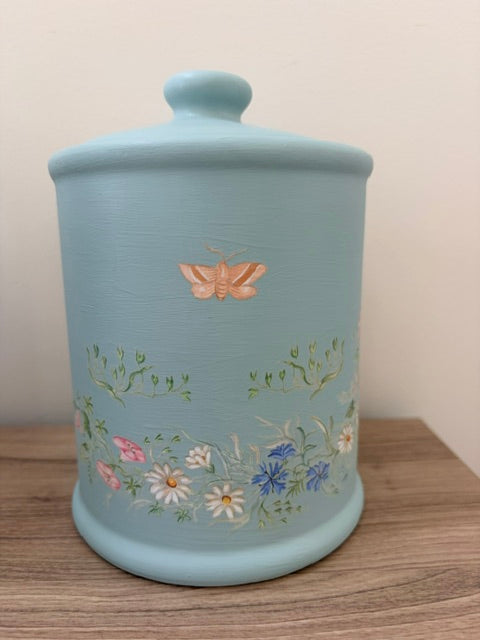 A light blue ceramic canister with a matching lid, decorated with a gold butterfly and a band of colorful hand-painted wildflowers, sitting on a wooden surface against a plain wall.