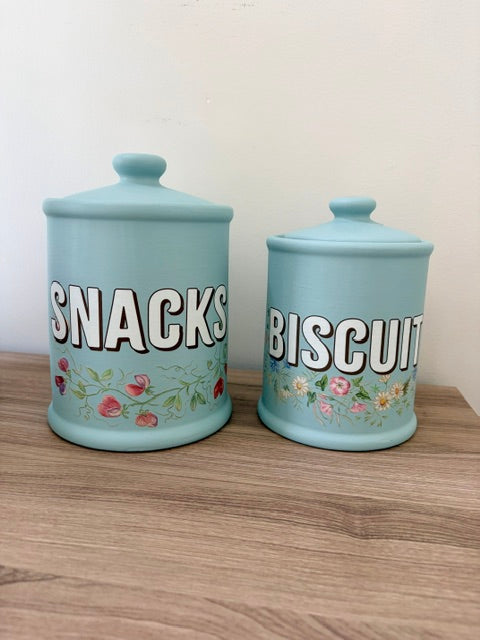 Two light blue ceramic canisters with lids, one labeled 'SNACKS' and the other 'BISCUIT', decorated with small floral illustrations, sitting on a wooden surface against a plain wall.