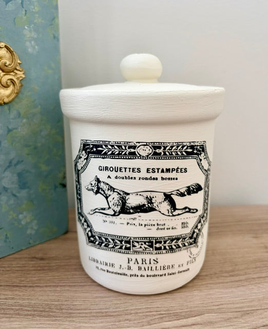 A white ceramic jar candle with a lid, featuring a vintage-style black label that reads 'GIROUETTES ESTAMPÉES PARIS'. The candle is placed on a wooden surface next to a blue-green decorative box.