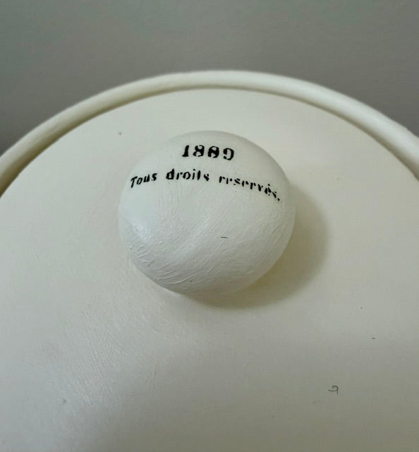 A close-up of a white ceramic or porcelain container lid with a round knob featuring black printed text that reads '1899' and 'Tous droits reservés.'.