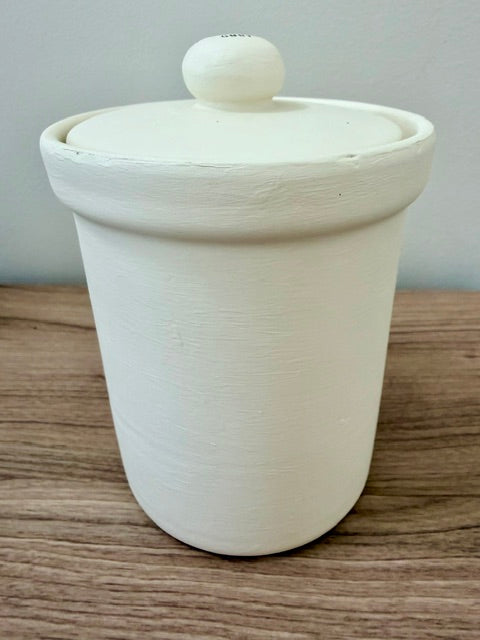 An off-white ceramic lidded container with a rounded cylindrical body and a small round knob on top, sitting on a wooden surface against a plain light gray wall.