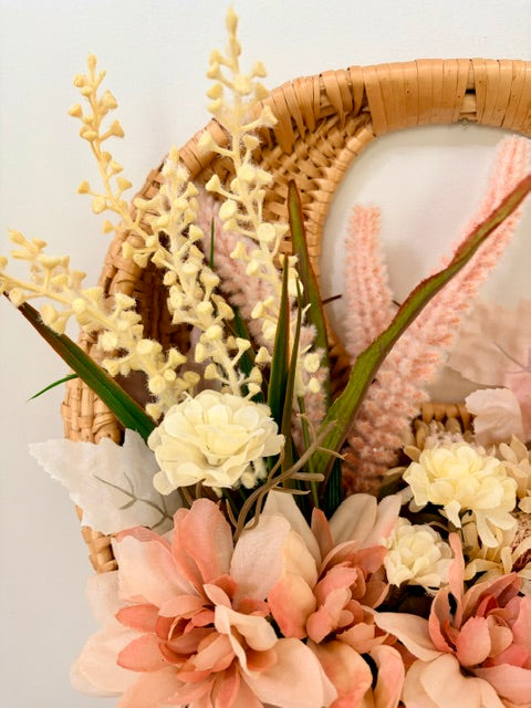 A close-up of a woven wicker basket filled with artificial flowers and greenery, including cream blossoms, peach and blush roses, and pale pink pampas-style stems, against a plain light background.