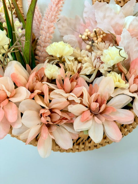 A close-up of a woven basket filled with artificial floral decor in soft blush pink, peach, cream, and pale yellow tones, with green stems and dried grass accents against a light blue background.