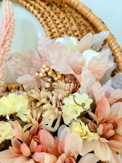A close-up of a decorative floral arrangement featuring large blush pink, cream, and peach flowers with small beige berries, styled in a wicker basket against a pale blue background.