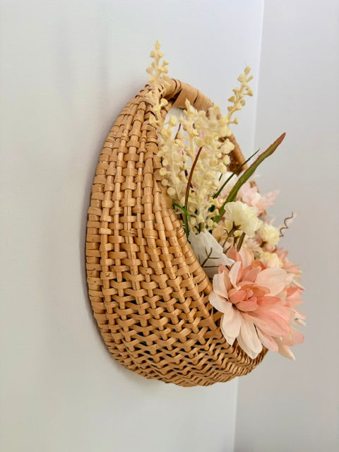 A natural woven wicker wall basket with a curved shape, mounted on a white wall and filled with artificial cream and pink flowers.