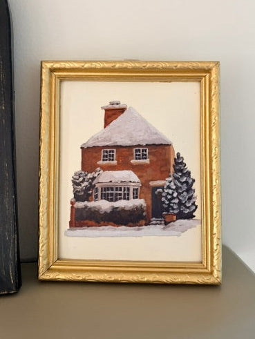 A framed watercolor painting of a snow-covered brick house, displayed on a shelf against a white wall.
