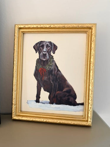 A framed portrait painting of a black Labrador retriever wearing a green wreath and red bow, sitting in white snow, displayed in a gold frame on a shelf against a white wall.