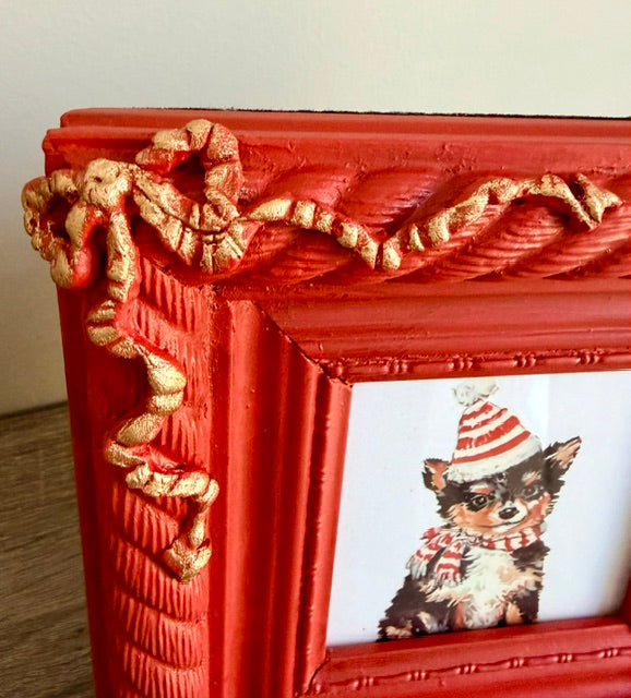 A close-up of a red picture frame with ornate gold rope-like corner decorations and a sample photo of a small dog wearing a red-and-white striped hat and scarf.
