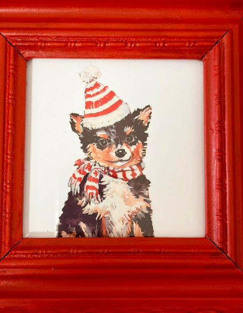 A framed painting of a small black and white dog wearing a red and white striped Santa hat and matching striped scarf, set within a bright red painted frame.