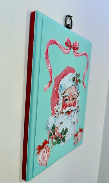 A vertical rectangular Christmas wall hanging with a turquoise background, featuring a painted Santa Claus face, pink ribbon, holly, and ornaments, suspended by a black metal ring against a white wall.