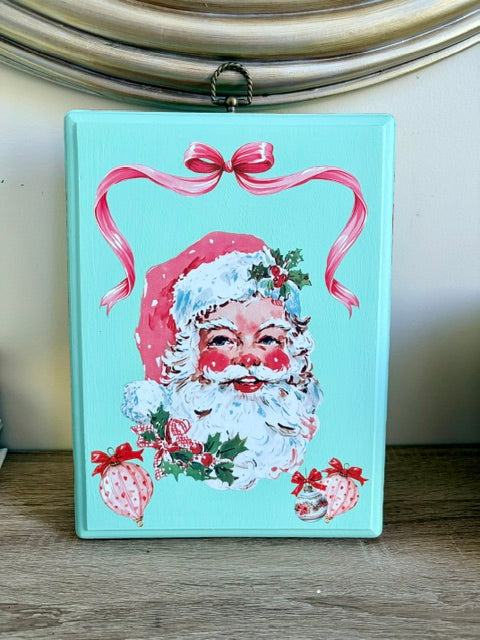 A rectangular turquoise Christmas ornament or plaque featuring a vintage-style Santa Claus face with a pink bow above, holly accents, and hanging loops at the top, displayed on a wooden surface.