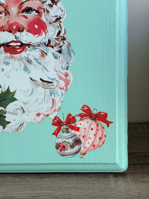 A close-up of a painted Christmas canvas or box corner featuring a partial Santa face with white beard, red nose, and red lips on a mint background, with two red-bow ornaments near the bottom right.