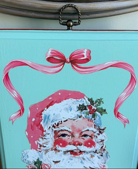 A close-up of a Christmas-themed decorative sign or box panel in light aqua, featuring a painted Santa Claus face with a red hat, white beard, pink ribbon above, and a hanging metal loop at the top.