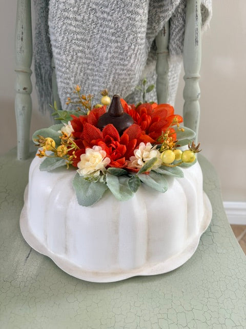 A white ceramic pumpkin-shaped centerpiece decorated with orange and red faux flowers, green leaves, and small yellow berries, displayed on a light green table with a gray chair and blanket in the background.