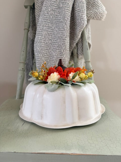 A white fluted metal pan decorated with a fall floral arrangement of red, orange, yellow, and cream flowers with green leaves, styled on a rustic table with a gray textured throw blanket draped over a distressed green chair.