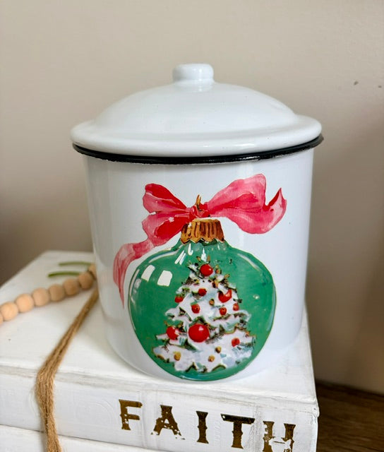 A white enamel-style lidded jar with a black rim, decorated with a painted green Christmas ornament featuring a white tree, red ornaments, and a pink bow. The jar is styled on stacked white books with gold lettering.