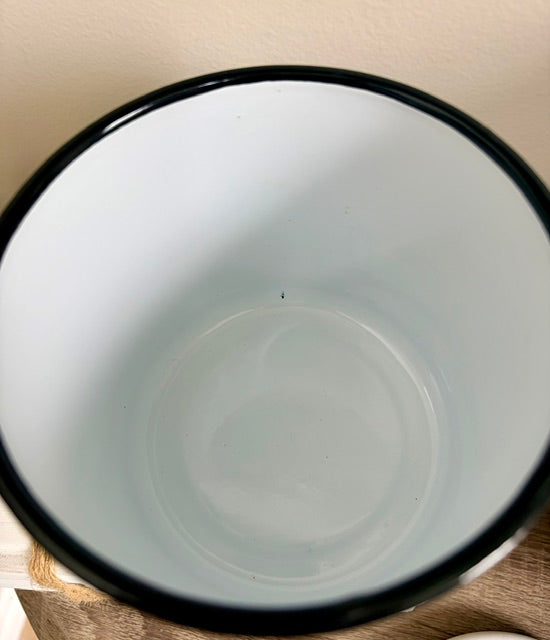 A top-down view of a white enamel bowl with a black rim, resting on a wooden surface against a beige wall.