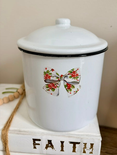 A white enamel lidded canister with a black rim and a decorative bow and holly design on the front, displayed on a white book labeled 'FAITH'.