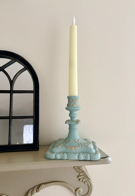 A light blue ornate candlestick holder with a cream taper candle, displayed on a shelf next to a black-framed mirror against a beige wall.