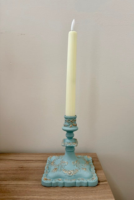 A decorative teal blue candlestick holder with gold floral accents holding a cream-colored taper candle, set on a wooden surface against a plain beige wall.
