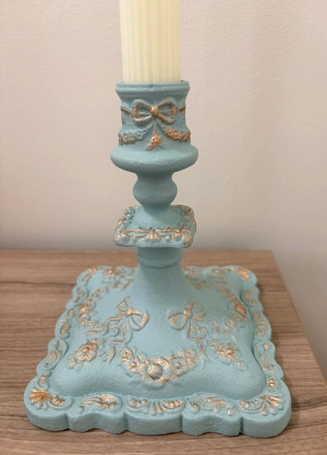 A decorative light blue candlestick holder with gold-painted floral and bow details, holding an off-white candle, set on a wooden surface against a neutral wall.