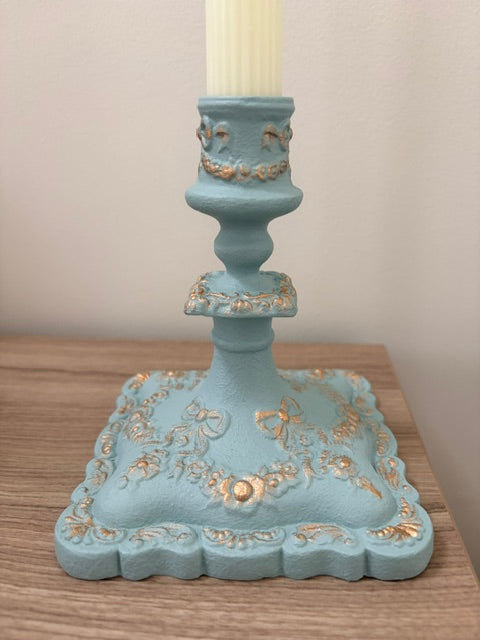 A decorative cast iron candle holder in a light blue finish with gold leaf accents, holding a cream-colored candle, set on a wooden surface against a neutral wall.