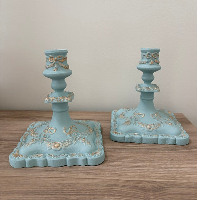 A pair of decorative light blue candlesticks with gold floral and bow accents, displayed on a wooden surface against a plain beige wall.