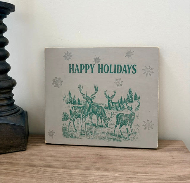 A rectangular decorative sign with a light gray background and teal holiday illustration of deer, reindeer, snowflakes, and the words “HAPPY HOLIDAYS,” displayed on a wooden surface against a white wall.