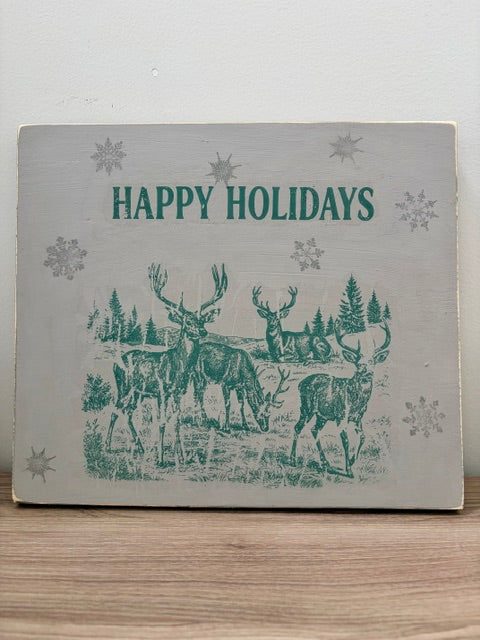 A square decorative sign with a distressed off-white background featuring a teal-green holiday illustration of deer in a forest and the words 'HAPPY HOLIDAYS' at the top, displayed on a wooden surface against a light wall.