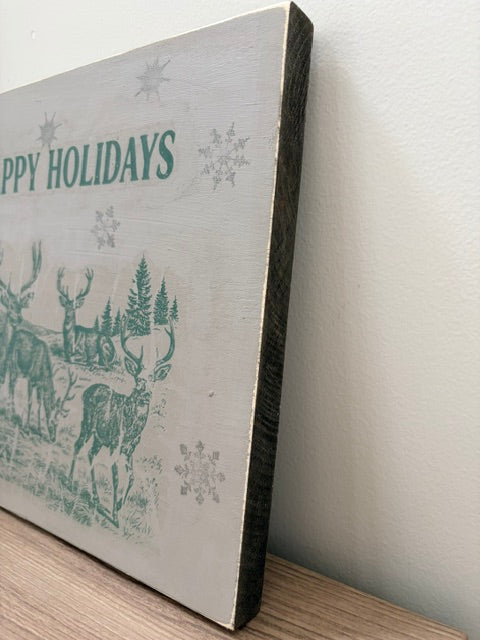 A rectangular decorative sign with a distressed off-white background, featuring green text reading 'HAPPY HOLIDAYS' and a green winter scene with deer, trees, and snowflakes, leaning against a light wall on a wooden surface.