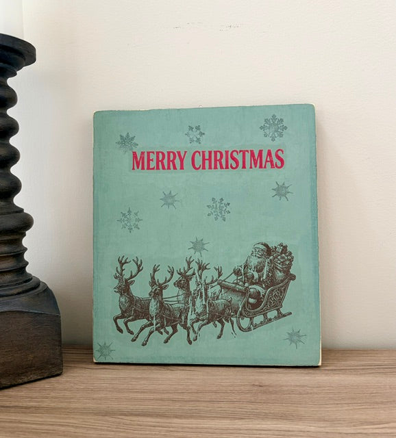 A vintage Christmas wood sign with a mint green background, red 'MERRY CHRISTMAS' text, and a dark illustration of Santa in a sleigh with reindeer, standing on a wooden surface against a light wall.