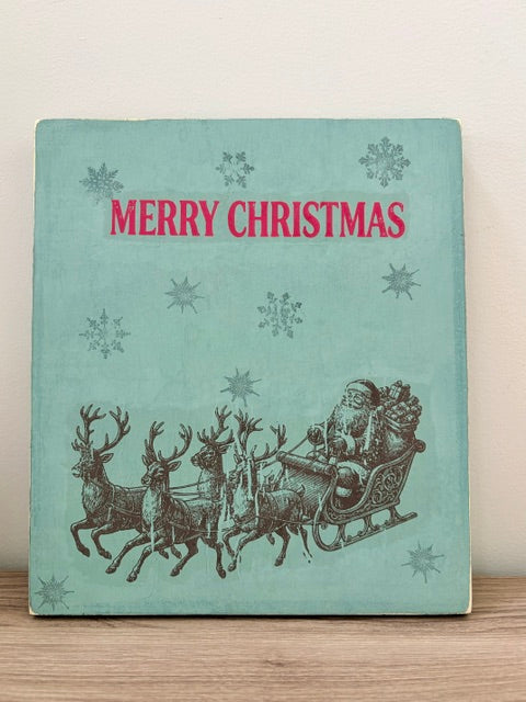 A square decorative sign with a vintage Christmas design, featuring the words 'MERRY CHRISTMAS' in red at the top and an illustration of Santa Claus in a sleigh pulled by reindeer at the bottom. The background is a light teal color with snowflake motifs.