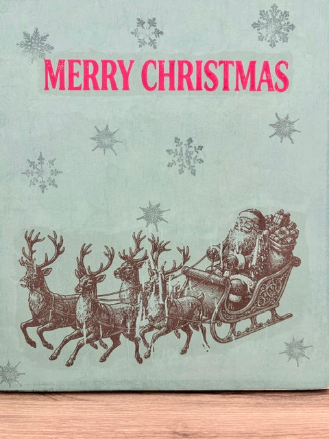 A vintage-style Christmas print or card with the words “MERRY CHRISTMAS” in bright pink at the top, a pale mint-green background with gray snowflakes, and a sepia-toned illustration of Santa Claus in a sleigh pulled by four reindeer near the bottom. The item appears photographed against a wood surface.