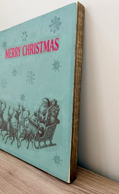 A square wooden Christmas wall sign with a light blue-green background, featuring a vintage illustration of Santa Claus in a sleigh with reindeer and the words 'MERRY CHRISTMAS' in pink. The sign is leaning against a white wall on a wooden surface.