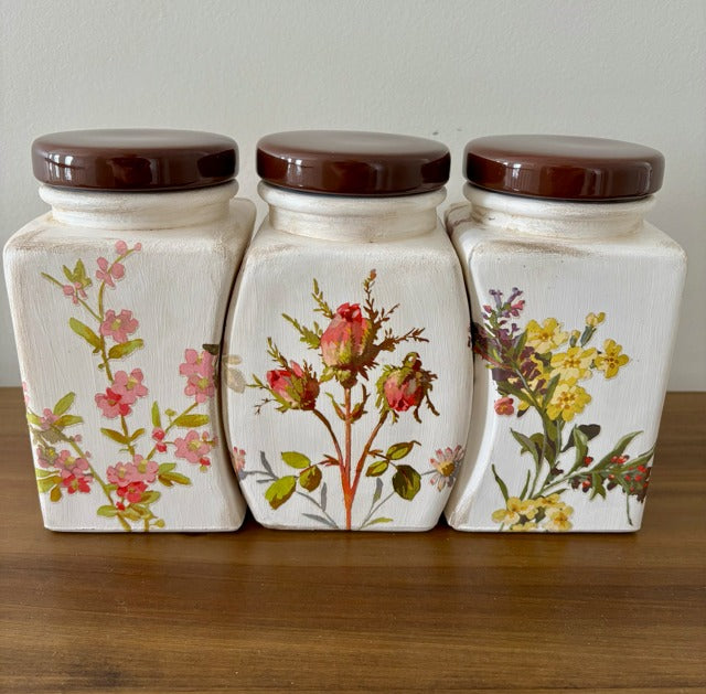 Three ceramic jars with floral designs and brown lids on a wooden surface.