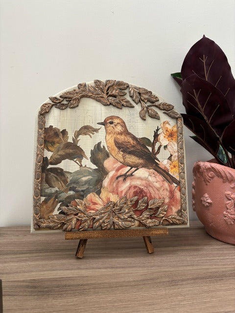 Decorative piece with a bird and floral design on a wooden stand, placed on a surface with a plant in the background.