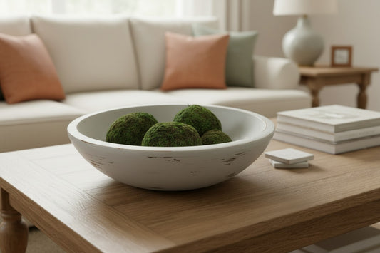 White bowl with green decorative balls on a wooden coffee table in a living room.