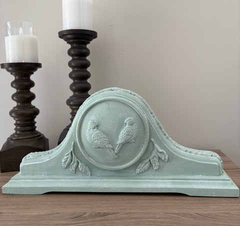Decorative wall art piece with bird design on a wooden surface with candles in the background.