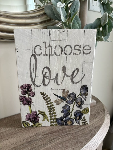 Decorative sign with 'choose love' text and floral designs on a wooden surface.