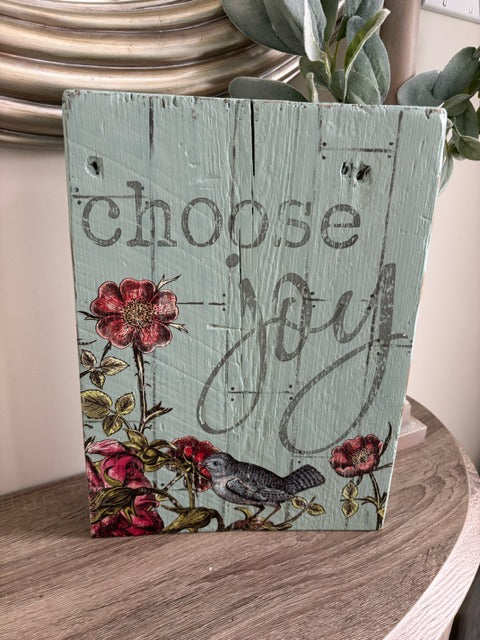 Decorative wooden sign with floral design and 'choose joy' text on a wooden surface.