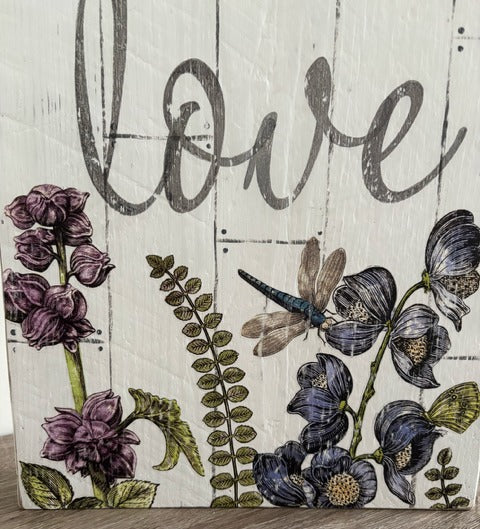 Decorative sign with flowers, a dragonfly, and the word 'love' on a wooden background.