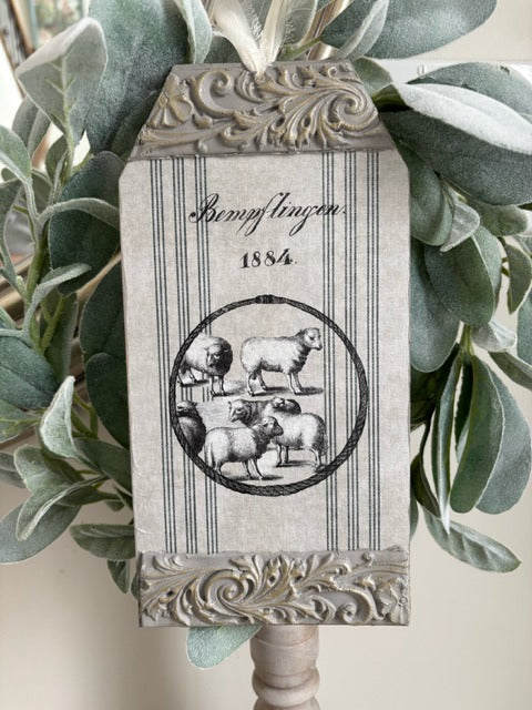 Decorative sign with sheep illustration and text on a stand with green leaves in the background