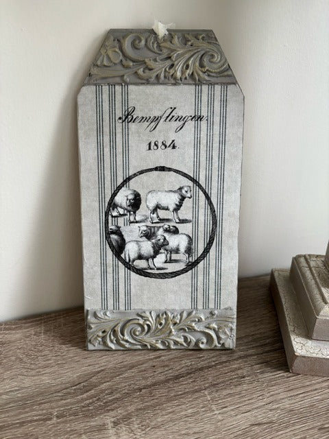 Decorative wall hanging with sheep illustration and text on a wooden surface.