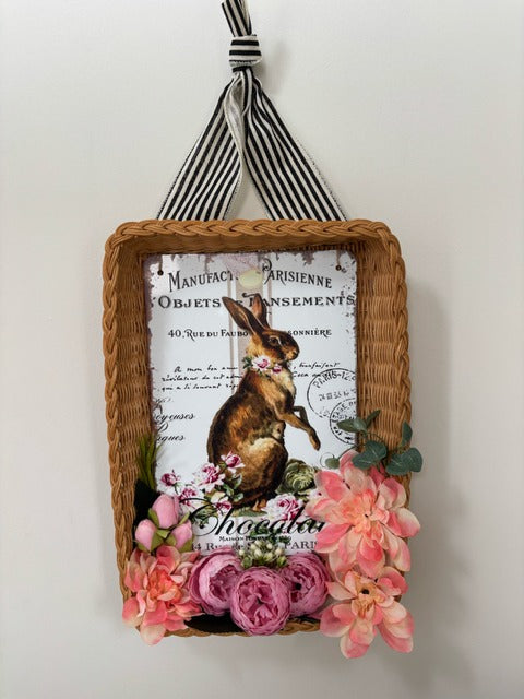 Decorative wall hanging with a rabbit illustration and floral elements, framed in wicker.